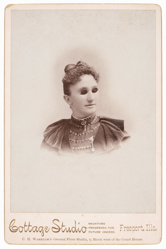 A black-and-white studio photograph of the head and shoulders of a White woman in Victorian-style dress wearing small, dark glasses.