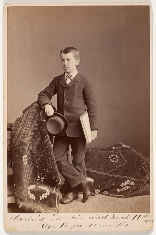 A sepia-toned photograph of a White boy in a suit, standing on a draped carpet, holding a notebook under one arm and a felt hat in his hand.