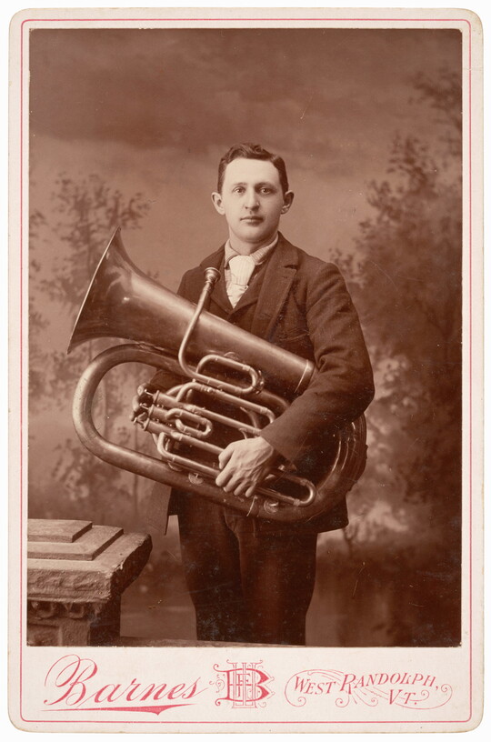 A black-and-white studio photograph of a White man in a suit holding a large trumpet-like brass instrument against his torso.