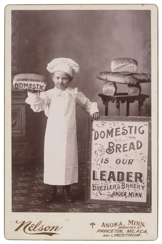 A black-and-white studio photograph of a White child dressed as a baker, holding up a loaf of bread that says "DOMESTIC" on its side, and standing next to a table of bread and a sign that reads, "Domestic bread is our leader, Brezler's Bakery, Anoka, Minn."