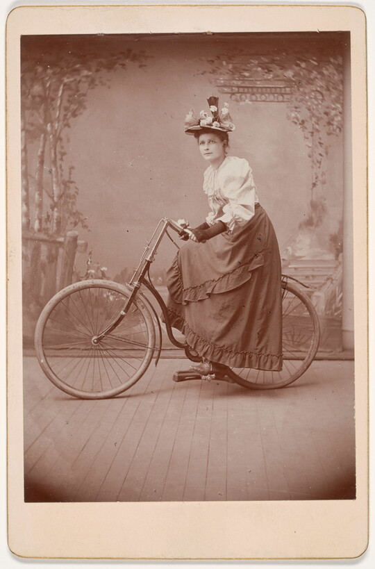 A black-and-white studio photograph of a White woman wearing a floral hat, white blouse, and long skirt posing on a bicycle in front of a scenic backdrop.