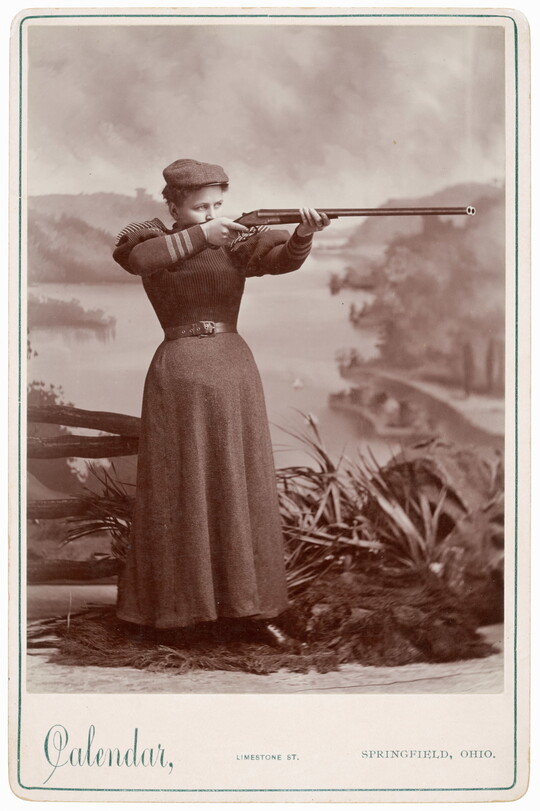 A black-and-white studio photograph of young White woman in a flat cap and plain, corseted gown aiming a rifle in front of a painted river scene.