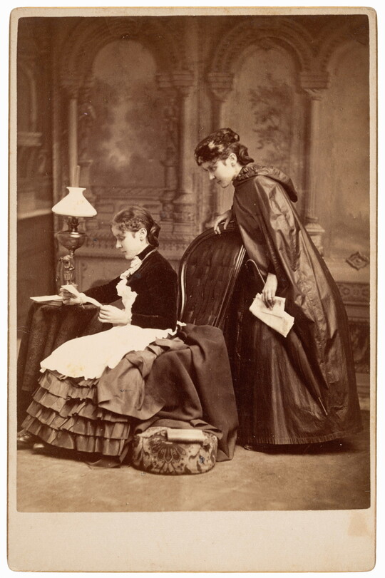 A black-and-white photograph of two identical White women in long dresses; one woman, seated, reads while the other stands behind and looks over her shoulder.