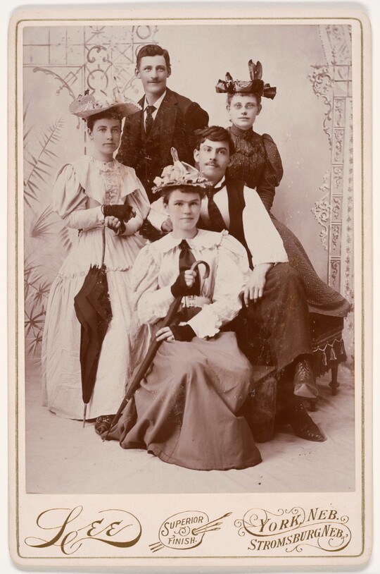 A black-and-white studio photograph of five White people: three women in long dresses and hats and two men, one in a suit, another in a vest.