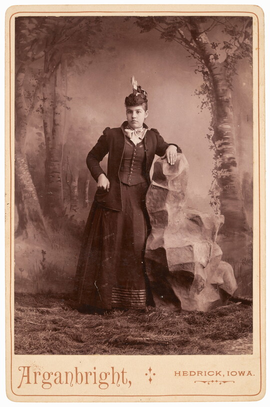 A sepia-toned studio photograph of a young White woman wearing a dark jacket, skirt and corseted vest, leaning on a fake rock in front of a painted forest landscape.