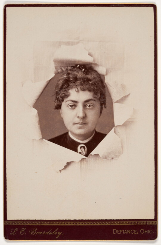 A black-and-white studio portrait photograph of a White woman behind paper that looks like it has been torn back to reveal her face.