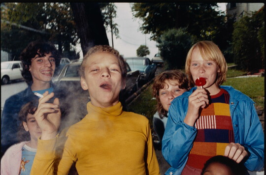 A color photograph of a group of smiling preteens: the boy in the front is smoking a cigarette and the one on the right is licking a lollipop.