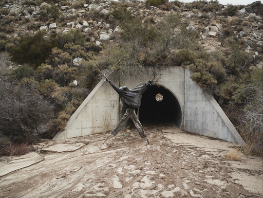 A color photograph of a human-like figure made from sticks and dressed in dark clothing in front of a drainage tunnel.