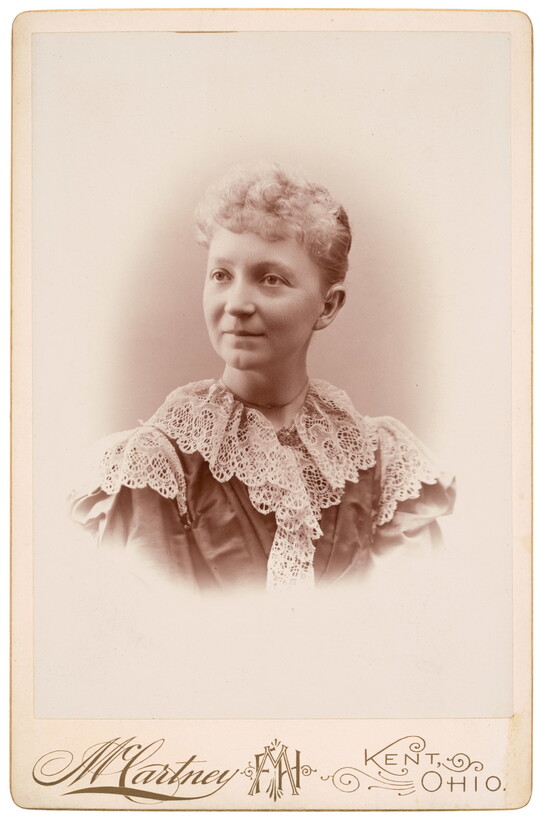 A sepia-toned studio portrait photograph of a White woman with curly, light colored hair in an updo, wearing a high-collared, frilly lace top.