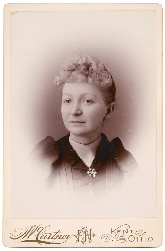 A sepia-toned studio portrait photograph of a White woman with curly, light colored hair in an updo, wearing a dark top closed with a brooch at the neck.