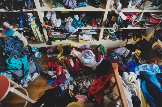 A color photograph of a light-skinned teenage girl lying on the floor of a messy room surrounded by unfolded clothing.