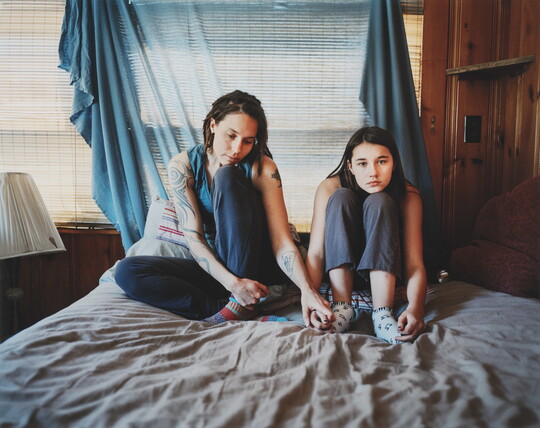 A color photograph of two light-skinned women holding hands and sitting on a bed in front of a fabric-covered window.