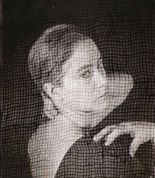 A black-and-white portrait photograph of a White woman, head turned to look over her shoulder, as viewed through what looks like a screen.