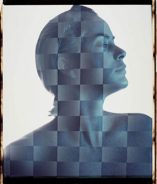 A blue-toned portrait photograph of a White woman with short hair, head turned in profile, as viewed through a checker-board like filter.
