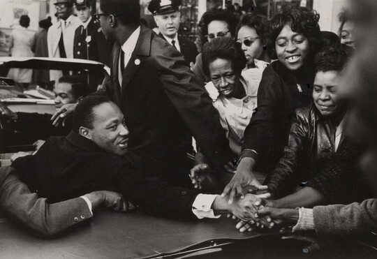 A black-and-white photograph of a Black man sitting in the back of a convertible and shaking hands with a crowd of Black people.