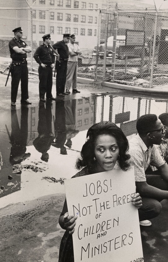 A black-and-white photograph of a Black woman holding a protest sign as White police officers stand in the background.