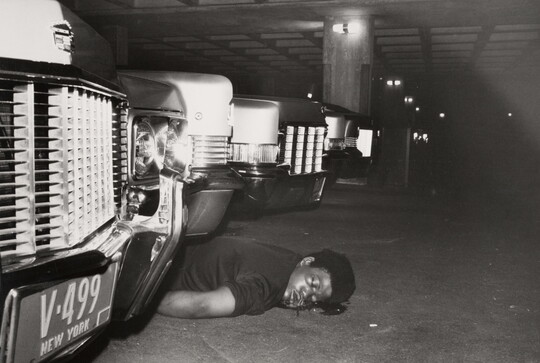 A black-and-white photograph of the body of a Black person laying on their stomach with a blood stain around their head between cars in a parking garage.