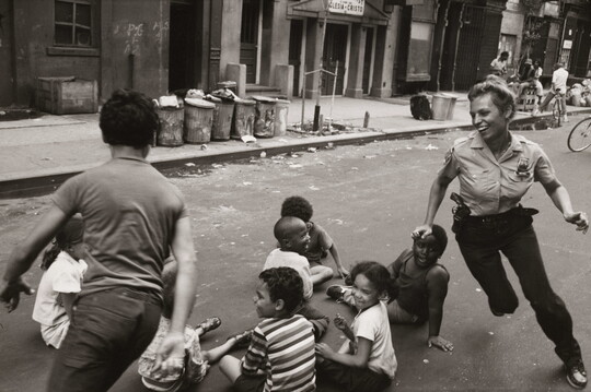 A black-and-white photograph of a smiling, White police woman playing a chase game with children of different races in a trash strewn street.