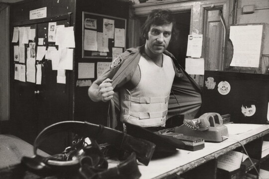A black-and-white photograph of a White man standing behind a cluttered counter holding open his shirt to show a bullet-proof vest.