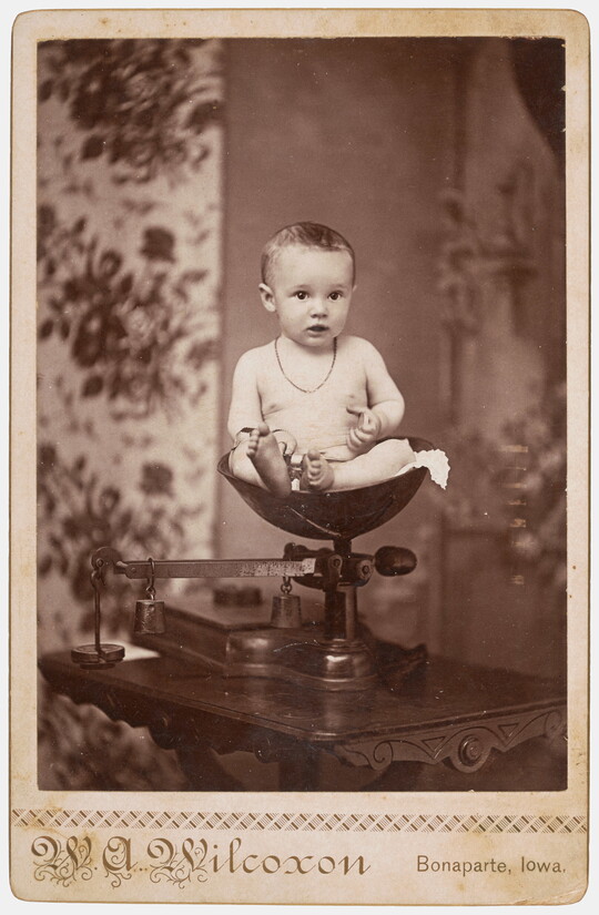 A sepia-toned studio photograph of a White baby wearing a necklace sitting in a scale.