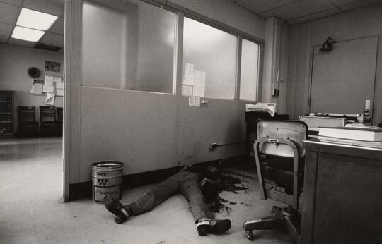 A black-and-white photograph of an office with a bloody body on the floor, feet toward the camera, the top part of the body obscured by dark stains on it, the floor, and the wall.