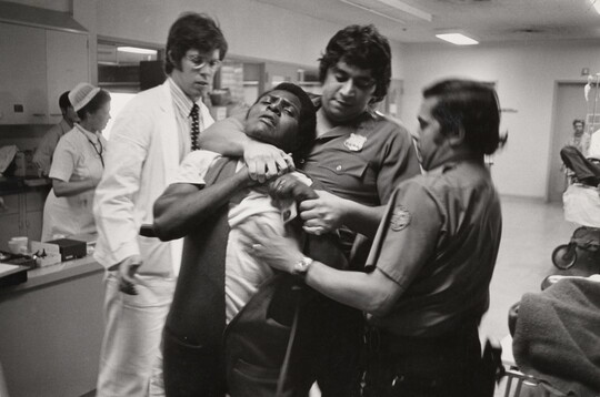 A black-and-white photograph of a Black man being restrained and choked by two police officers with medium to dark skin in a hospital setting.
