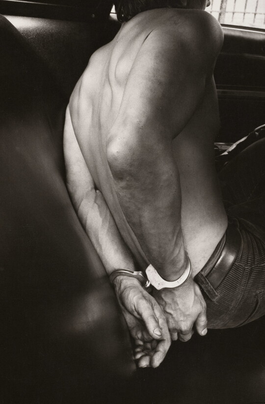 A black-and-white photograph of the bare back and handcuffed hands of a White man in the back seat of a police car.