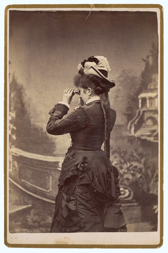 A sepia-toned studio photograph of a light-skinned woman, viewed from the back, wearing a dark corseted gown and peering through binoculars toward a painted landscape.