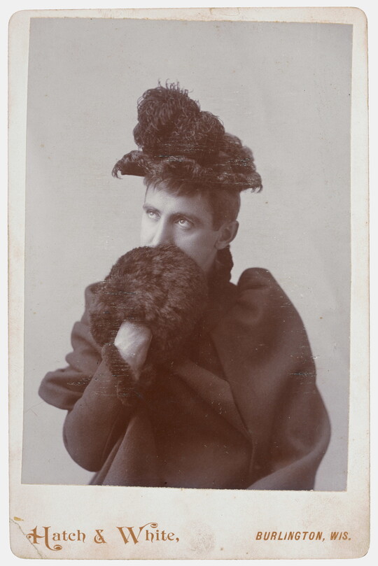 A black-and-white studio portrait photograph of a White person wearing a feather-trimmed hat, dark coat, and a fur muff that they are holding up in front of their mouth.