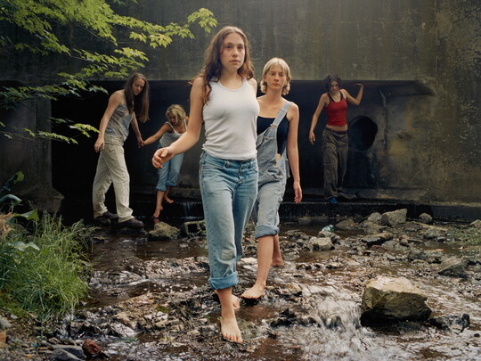 A color photograph of White teenagers, casually dressed, several barefoot, walking through a stream that emerges from a culvert behind them.