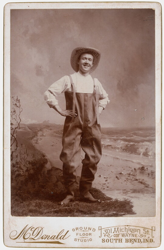A sepia-toned studio photograph of a smiling White man wearing waders and a large straw hat, posed with hands on hips.