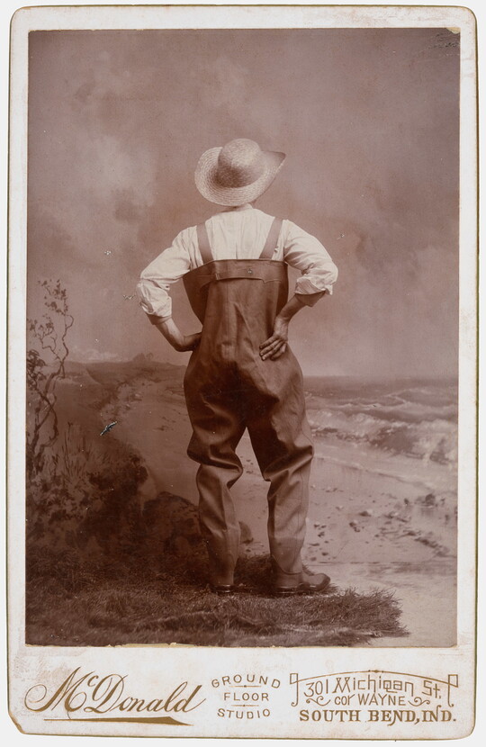 A sepia-toned studio photograph of a man, viewed from the back, wearing waders and a large straw hat, posed with hands on hips.