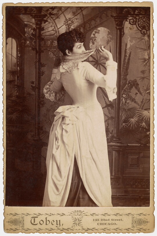 A sepia-toned studio photograph of a young White woman, viewed from the back, with a dramatically corseted dress and full bustle peeking playfully over her shoulder and hiding her face with a handkerchief.