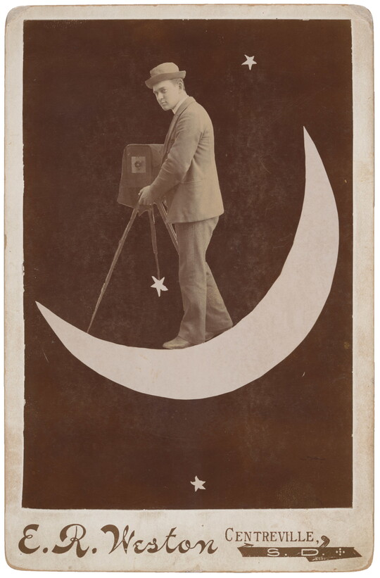 A black-and-white photograph of a White man wearing a bowler hat and suit holding an old-fashioned camera on a tripod while standing on a crescent moon.