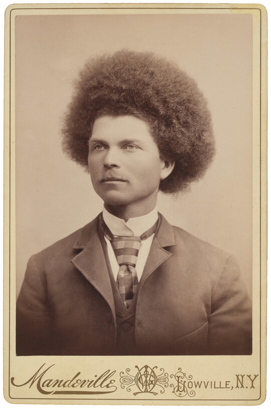 A sepia-toned studio portrait photograph of a man with "Afro"-style hair wearing a suit, collared shirt, vest, and tie.