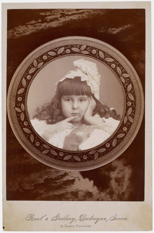 A black-and-white studio photograph of a young White girl with curled dark hair, bangs, and a massive bow on her head holding one finger to her mouth.