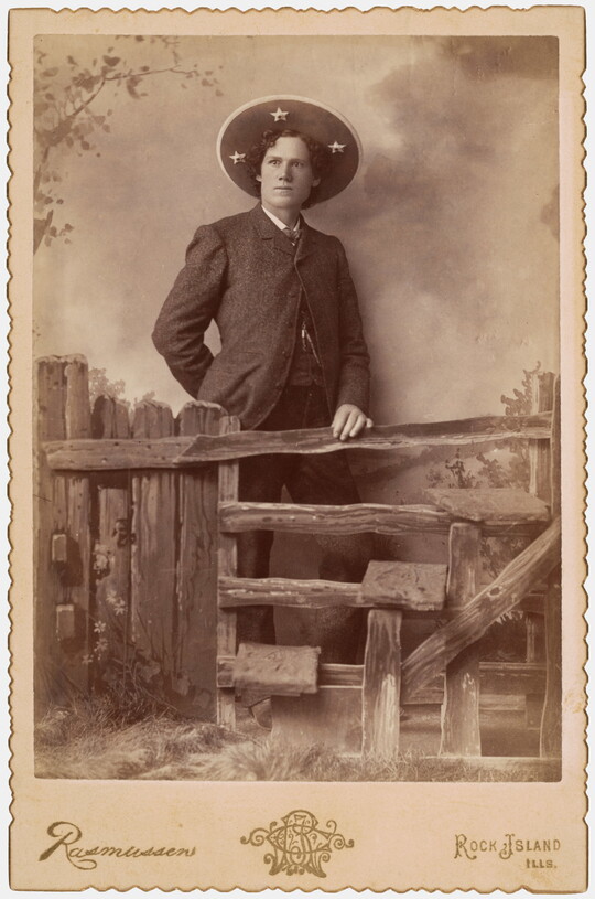 A sepia-toned photograph of a White man wearing a large sombrero-like hat standing behind a split-rail fence that is part of a studio set.