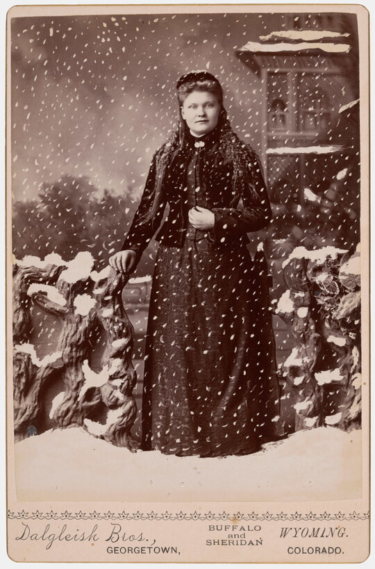 A black-and-white studio photograph of a White woman standing in a fake snowstorm against a backdrop of a winter scene.