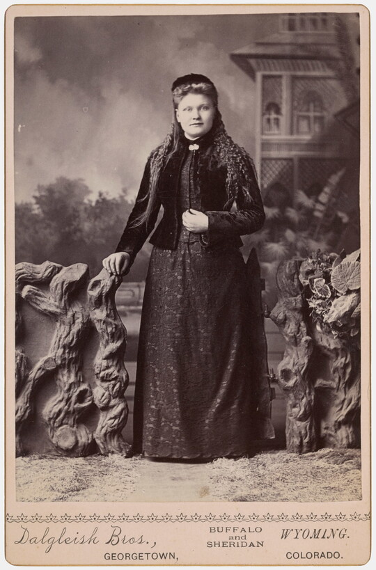A black-and-white studio photograph of a White women dressed in black standing in front of a backdrop of an outdoor scene.
