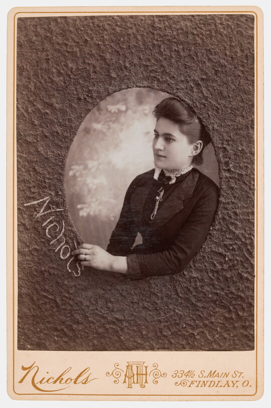 A sepia-toned studio photograph of a White woman who appears to be reaching through a textured oval window to write the word, "Nichols."