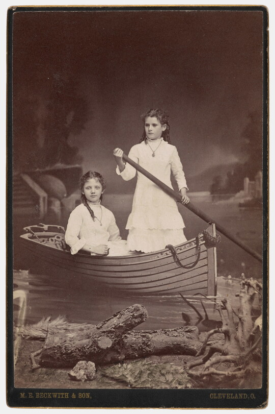 A sepia-toned studio photograph of two adolescent White girls, both wearing high-necked white dresses, posing in a rowboat in front of a painted lake scene.