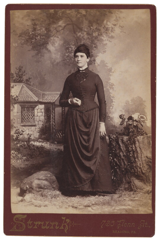 A sepia-toned studio photograph of a White woman with dark hair wearing a corseted, high-necked dress standing in front of a painted pastoral scene with a cottage.