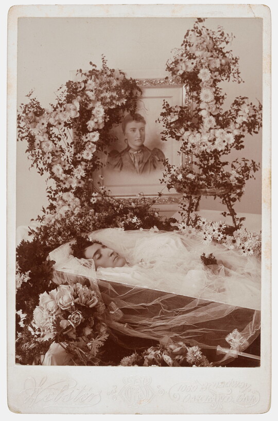 A sepia-toned photograph of a deceased woman in a coffin covered with flowers and a framed photograph of her in life on top of the coffin.