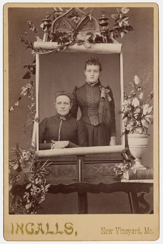 A sepia-toned studio portrait photograph of two White women on a massive paper scroll mounted on an easel and surrounded by flowers.