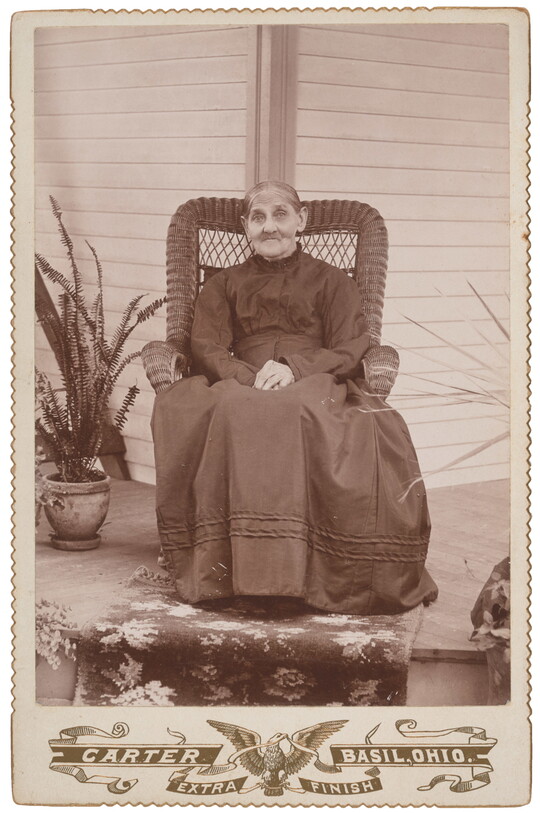 A sepia-toned studio photograph of an elderly White woman wearing a dark, high-necked dress sitting in a high back wicker chair on a porch with plants.