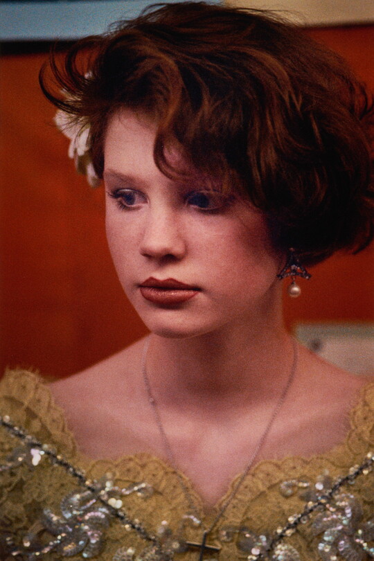 A color portrait photograph of a young White girl with short red hair, a flower tucked over one ear, and a decorative yellow top.