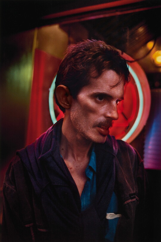 A color portrait photograph of a White man in profile with short brown hair, a mustache, prominent cheekbones, posed in front of neon lights.