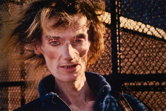 A color portrait photograph of a White person, short blond hair in disarray, prominent cheekbones and sunken cheeks covered in makeup, posed in front of a chain link fence.