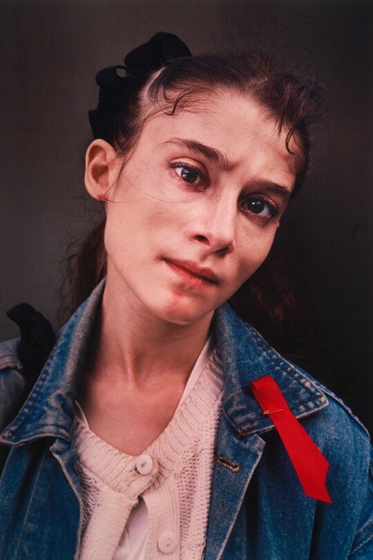 A color portrait photograph of a young fair-skinned woman with brown hair, wearing a light pink cardigan under jean jacket with a red ribbon pinned to the collar.