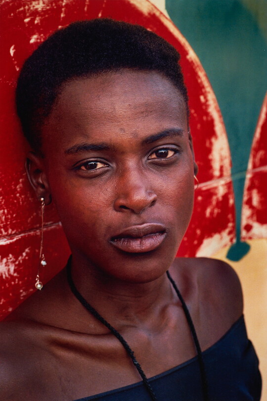 A color portrait photograph of a young Black woman with short hair, the straps of a black halter top up around her neck, posed against a red and green wall.